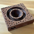 A square maple wood ravioli mold with a central circular shape and small holes on the surface, accompanied by a mini pin for pressing.