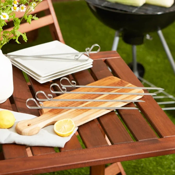 A set of six flat stainless steel skewers with looped ends, displayed on a wooden table next to a bbq.