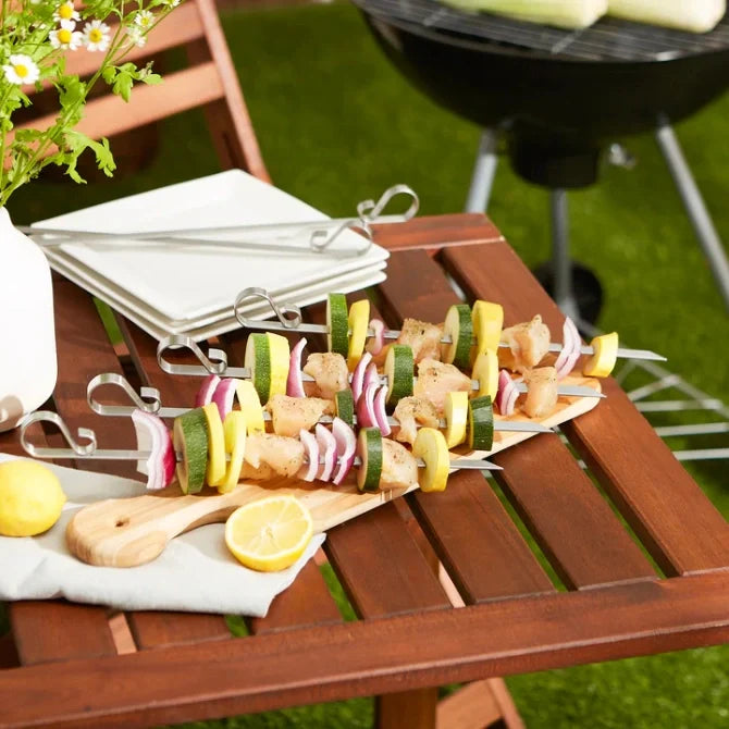 A set of six flat stainless steel skewers with looped ends, displayed against wooden table next to a bbq grill.