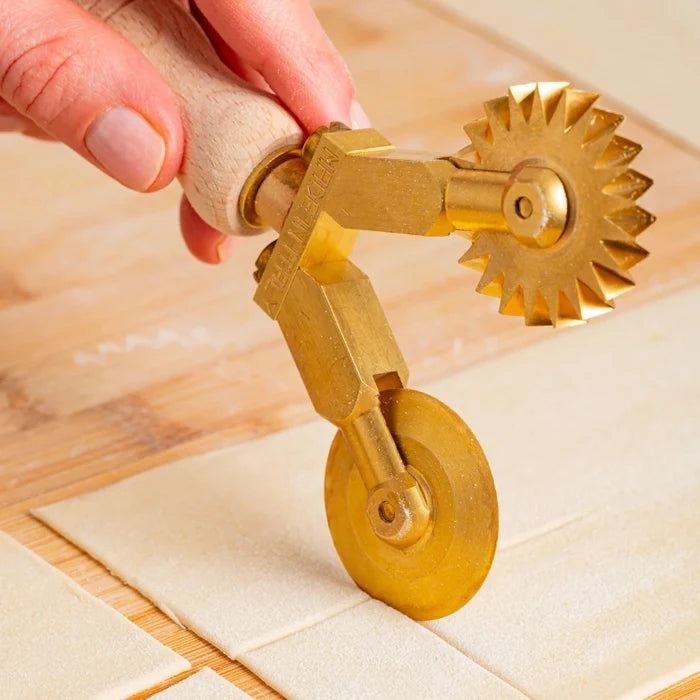 A handmade brass pasta cutter with a smooth and fluted wheel, featuring a wooden handle cutting out squares of dough.
